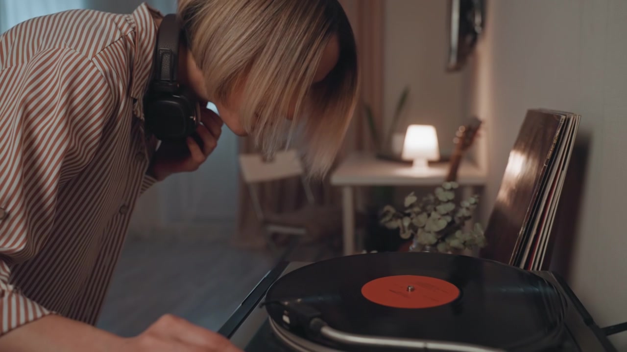 Donna caucasica amante della musica si china sul giradischi, posizionando attentamente il disco con le cuffie. Luce soffusa e accogliente nella stanza, primi piani delle mani che regolano il braccio e il piatto, un rituale audio concentrato e tattile.