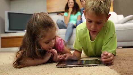 sibings lying on floor using tablet with parents behind them on sofa