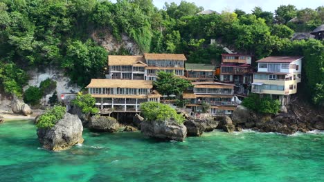 Luftdrohne-Schwenkt-über-Ein-Hotel-Und-Häuser-Auf-Einer-Klippe-In-Uluwatu,-Bali,-Während-An-Einem-Sonnigen-Tag-Eine-Indonesische-Flagge-über-Dem-Tropischen-Türkisblauen-Ozean-Weht