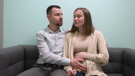 front view of pregnant woman and her husband sitting on sofa talking and waiting for gynecologist in medical consultation