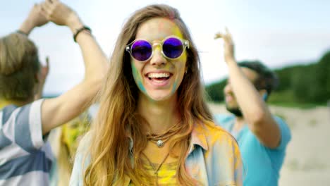 close-up portrait of a beautiful young girl with sunglasses standing in the crowd of people celebrating holi festival. people throwing colorful powder in her back. her face and clothes are covered with colorful powder.