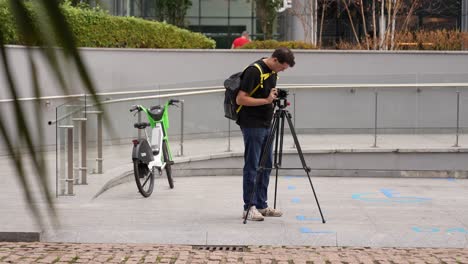 young man is standing taking photos and videos with his camera on a tripod next to an electric rental bike