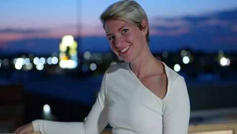 Beautiful-happy-short-blonde-haired-caucasian-women-in-white-dress-looking-and-smiling-into-camera-with-pink-and-blue-fading-sunset-and-city-lights