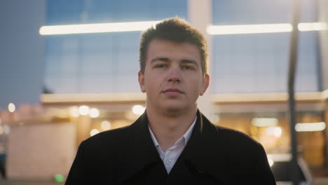 mall owner in black coat standing in front of mall with serious look, background features glowing street light, surrounding glow light, and glass building reflecting structures