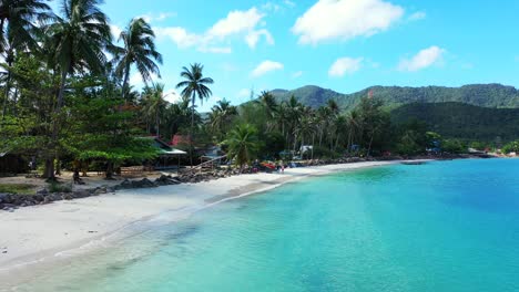 Ruhige-Bucht-Mit-Kristallklarem,-Smaragdgrünem-Wasser,-Das-Weißen-Sandstrand-Unter-Schatten-Von-Palmen-Am-Hellen-Himmel-Mit-Weißem-Wolkenhintergrund-In-Thailand-Wäscht