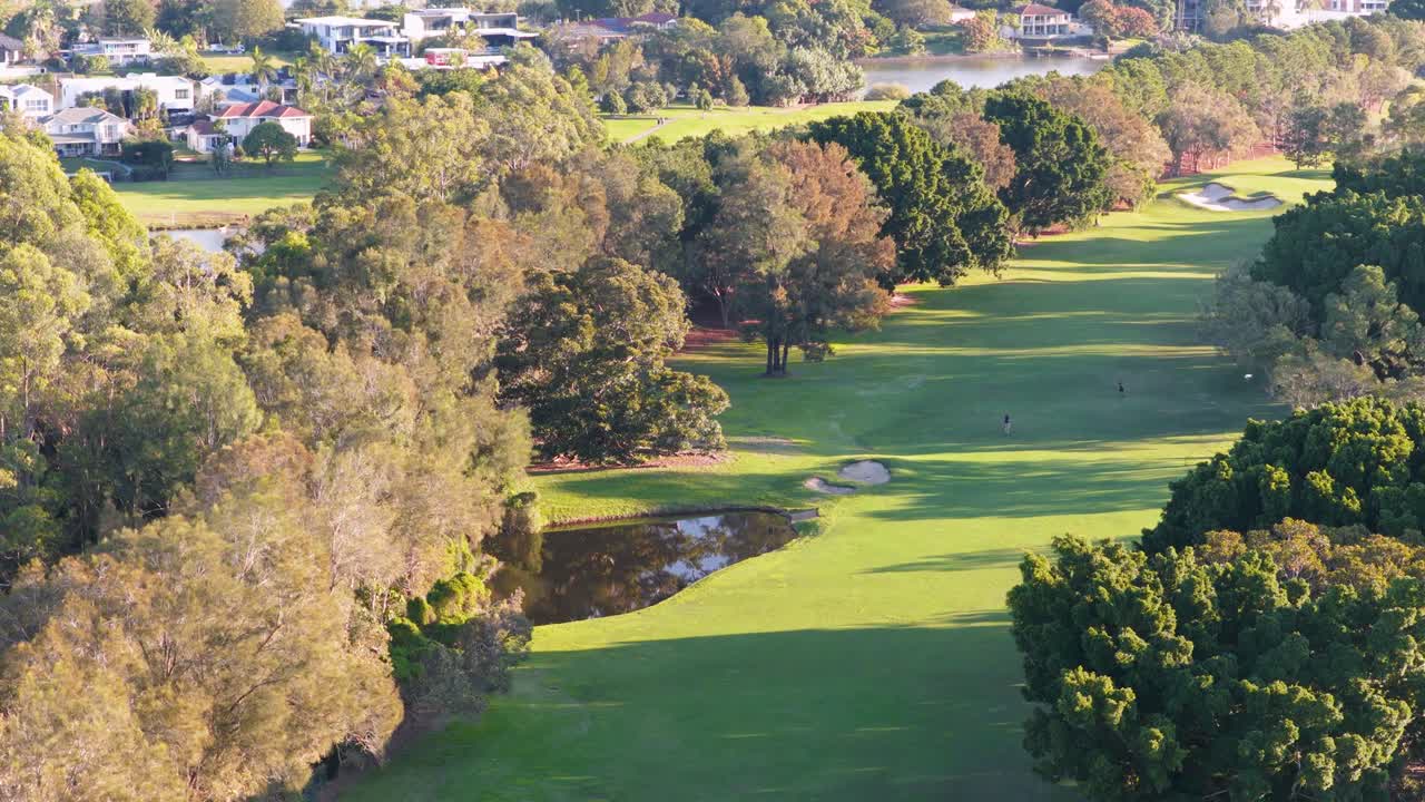 Drone footage captures a serene golf course landscape with vibrant greens, trees, and water hazards under clear skies