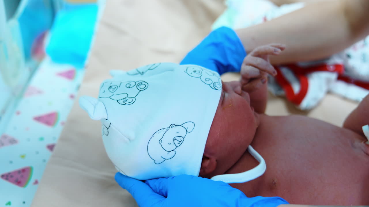 White cap with bears is put on the newborn. Gloved hands of medic tie the straps of cap while baby cries. Close up.