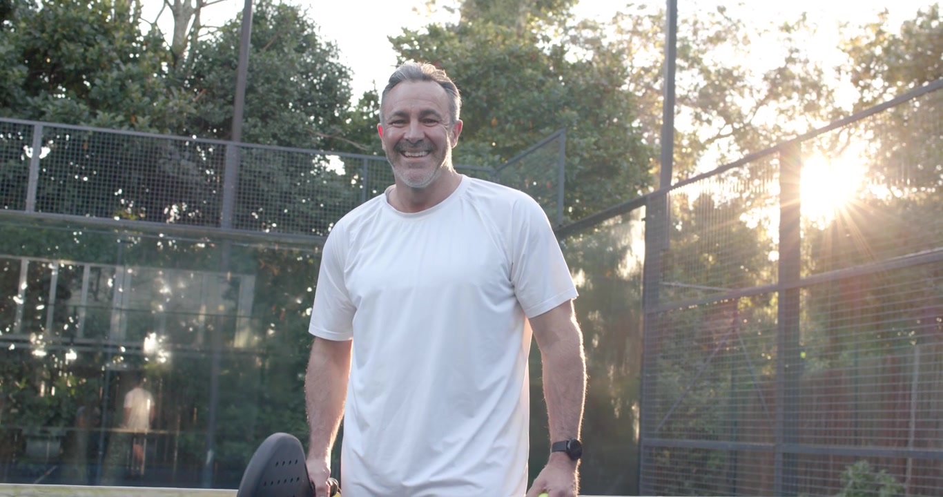 Hombre de mediana edad probando una pala con una pelota amarilla, caminando hacia la cámara en una cancha de pádel y saludando al espectador.