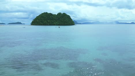 Drone-flies-over-ocean-waves,-sand-beach,-boats,-towards-a-small-green-island