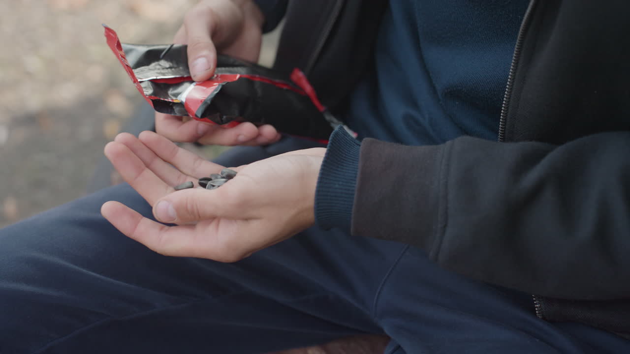 Uomo bianco seduto su una panchina del parco che apre una bustina di semi e ne versa il contenuto sul palmo delle mani, indossa un maglione blu e una giacca nera, primo piano sulle mani che condividono delicatamente i semi, atmosfera diurna tranquilla, interazione compassionevole