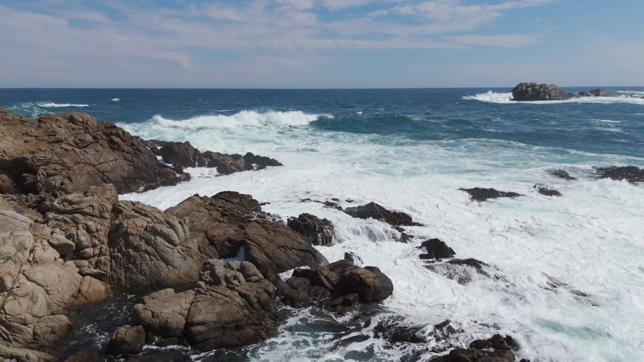 Aerial view of powerful ocean waves crashing against rugged coastal rocks, under a bright blue sky, symbolizing the untamed beauty and strength of nature at its finest.