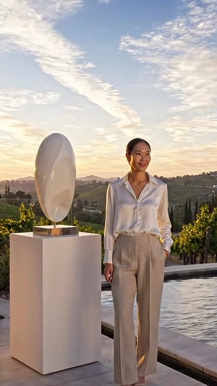Una mujer elegante con atuendo sofisticado posa junto a una escultura moderna bajo un cielo pintoresco, mostrando belleza y estilo en un entorno exterior tranquilo.