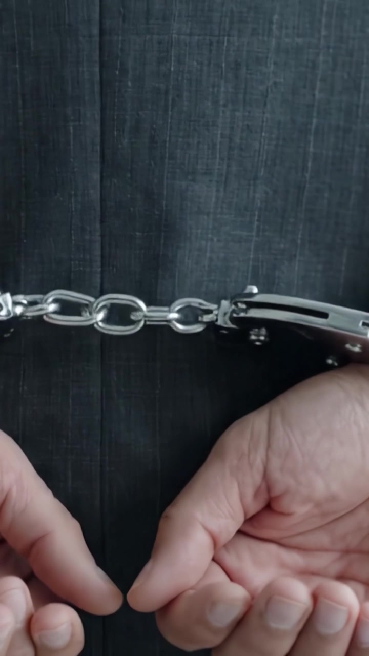 Una vista ravvicinata di manette assicurate ai polsi di una persona, che mostra un intenso momento di confinamento e restrizione, illustrato da mani strettamente legate e maglie di catena metalliche.