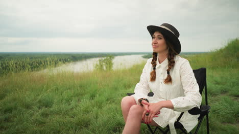 a beautiful woman wearing a hat and a white dress is seated on a chair in a lush grass field beside a tranquil lake, posing elegantly with a serene expression, under a cloudy sky