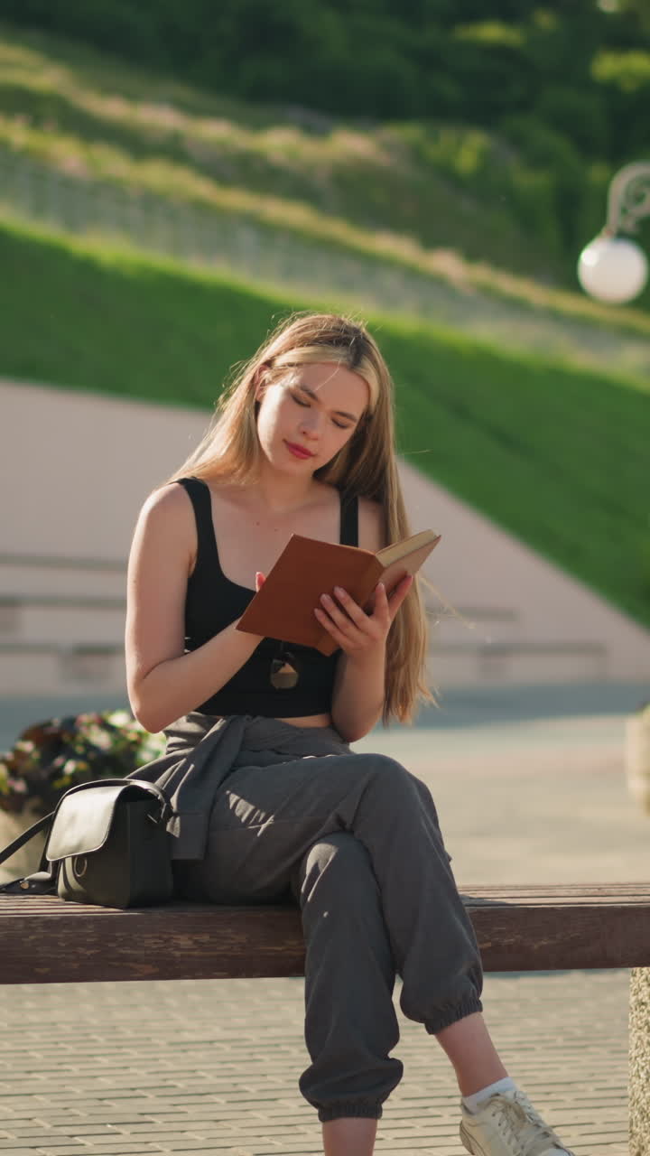 Woman seated outdoors on bench, reading a book thoughtfully with legs crossed, sun illuminating her, she holds the book gently, while the background features lined-up light poles
