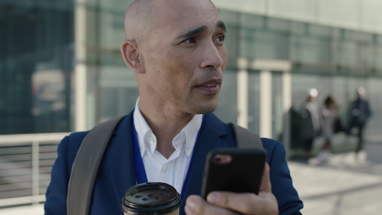 Nærbildeportrett av en skallet latinamerikansk forretningsmann som ser mot kamera, bruker smarttelefon og drikker kaffe på et kontor.