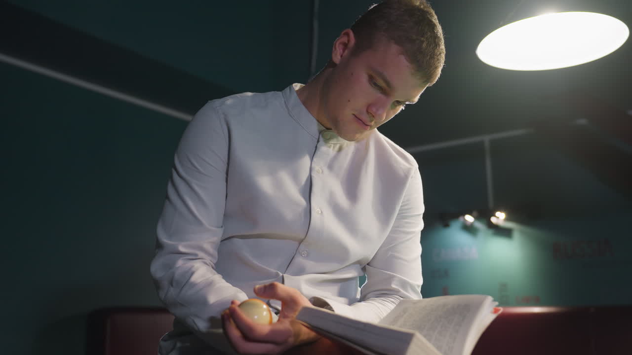 Close-up shot of dude in white shirt sitting on green pool table, reading book while rolling billiard ball in hand. Focused expression, dramatic lighting, moody background, and deep concentration
