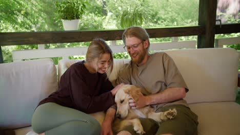 A-blond-girl-in-a-red-sweater-and-a-blond-guy-in-glasses-with-a-beard-in-a-light-brown-T-shirt-are-sitting-on-a-devan-and-stroking-their-big-light-colored-dog-on-a-devan-in-a-gazebo-in-nature