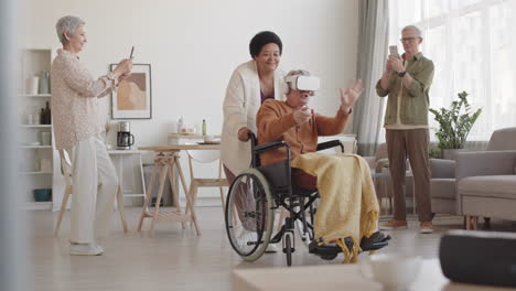 full shot of happy handicapped person wearing vr goggles with earphones, his female african friend moving wheelchair sideways, senior woman and man standing, filming them on smartphones