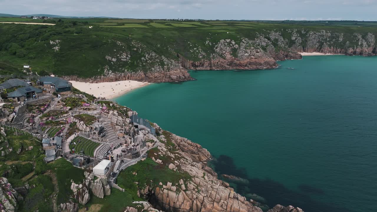 Rightward drone orbit shows Minack Theatre terraces and dramatic cliff edge by the sea, aerial establishing overview