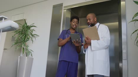 African-american-male-and-female-doctors-holding-clipboard-and-tablet,-talking-at-hospital