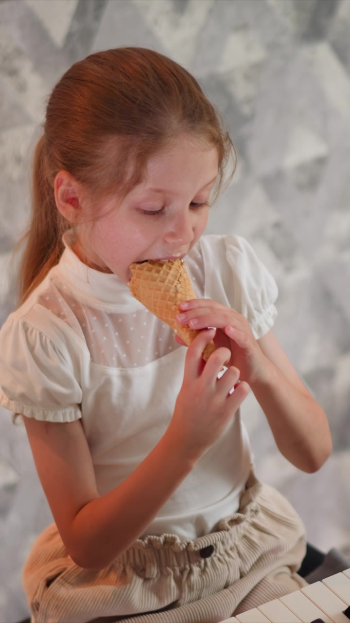 Close up of young girl eating ice cream cone while seated near piano keys. Girl wears white blouse with polka dot detail and beige corduroy pants holding waffle texture food item as girl