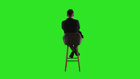 rear view studio shot of man sitting on stool facing away from camera against green screen