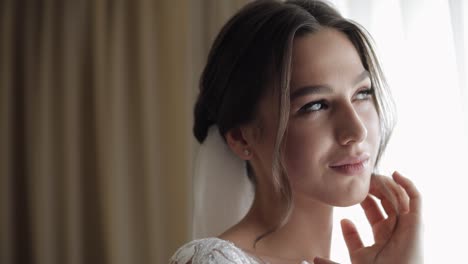 beautiful bride in white wedding dress smiles looking out window