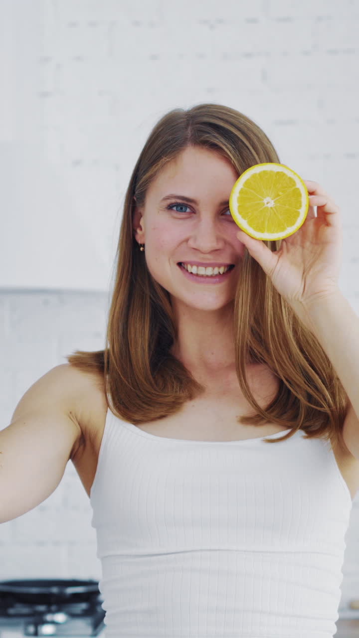 Beautiful woman holding two parts of orange on kitchen background. Happy female closing her eyes by cutted parts of orange indoors. Dieting concept. Vertical video