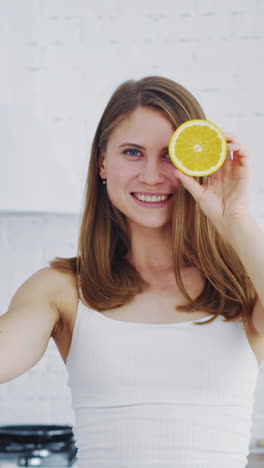 beautiful woman holding two parts of orange on kitchen background. happy female closing her eyes by cutted parts of orange indoors. dieting concept. vertical video