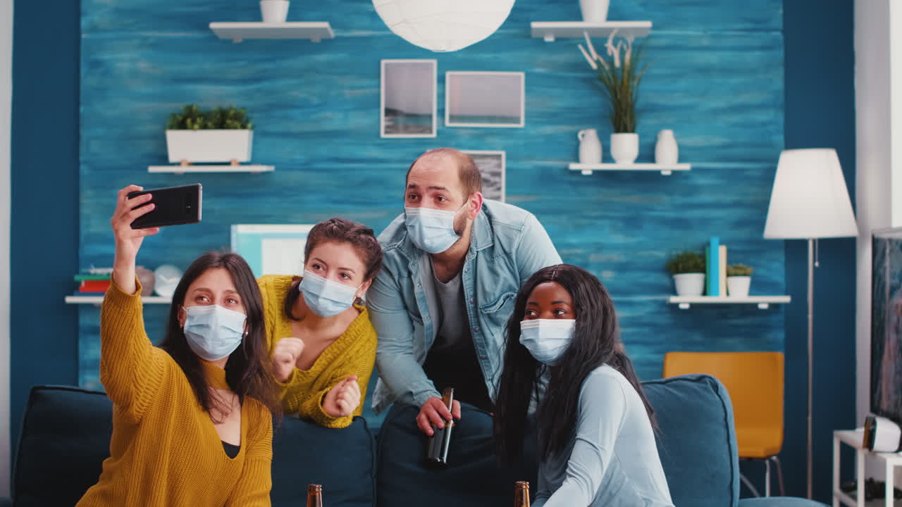 Multiracial friends sitting on sofa with protection masks, taking selfie