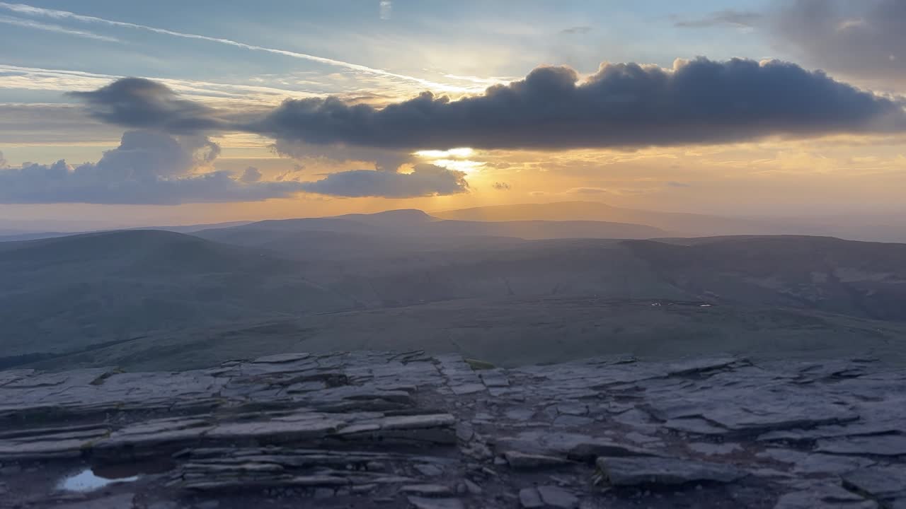 Rotsachtig bergplateau op Pen y Fan met dramatische wolken en zonsonderganglicht over verre glooiende toppen, wat een uitgestrekt berglandschap creëert met een sfeervolle avondstemming.