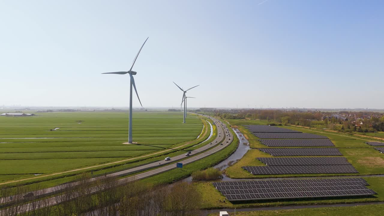 Wide aerial shot of Dutch countryside featuring renewable energy sources: tall wind turbines next to fields of solar panels along a modern highway. Captured in clear daylight.