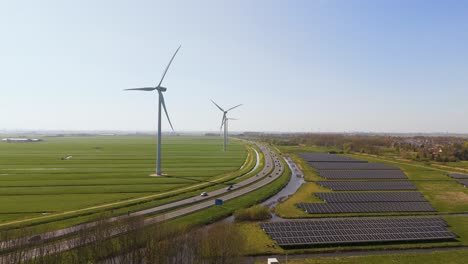wide aerial shot of dutch countryside featuring renewable energy sources: tall wind turbines next to fields of solar panels along a modern highway. captured in clear daylight.