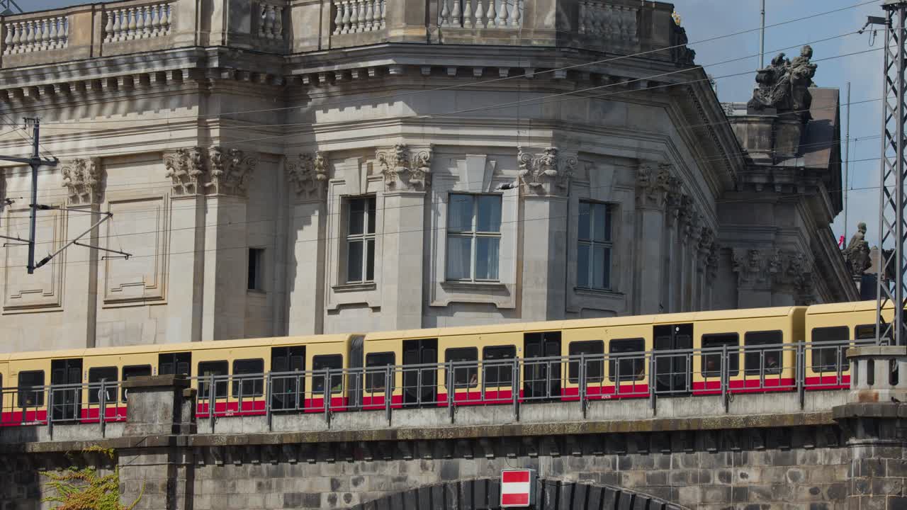 Yellow commuter train moves across arched stone bridge beside ornate classical building, daylight, static shot