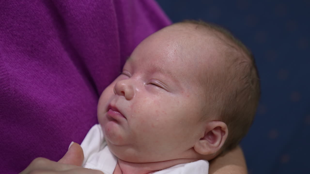 Sleepy newborn kid doesn't want to wake up lying in mum's hands. Mother is touching her baby's plump cheeks trying to wake him up. Close up.