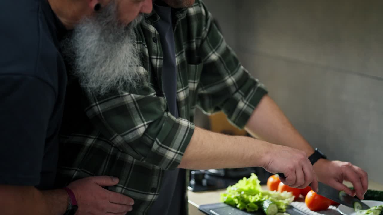 Close-up a middle-aged brunette man in a plaid shirt prepares a vegetable salad while his elderly boyfriend with gray hair and a bushy beard hugs the man and supports him in the kitchen before breakfast