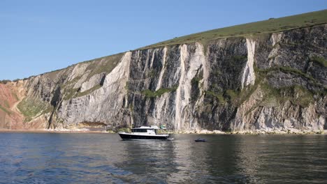 a boat anchored near tall, rugged cliffs with green patches under a clear blue sky