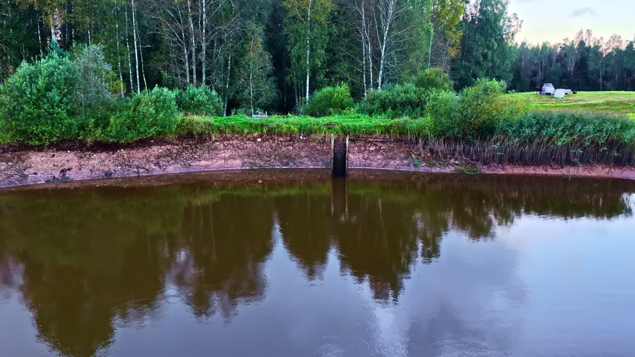 Drone zooms out revealing a wooden water gate along a muddy riverbank bordered by forest at sunrise