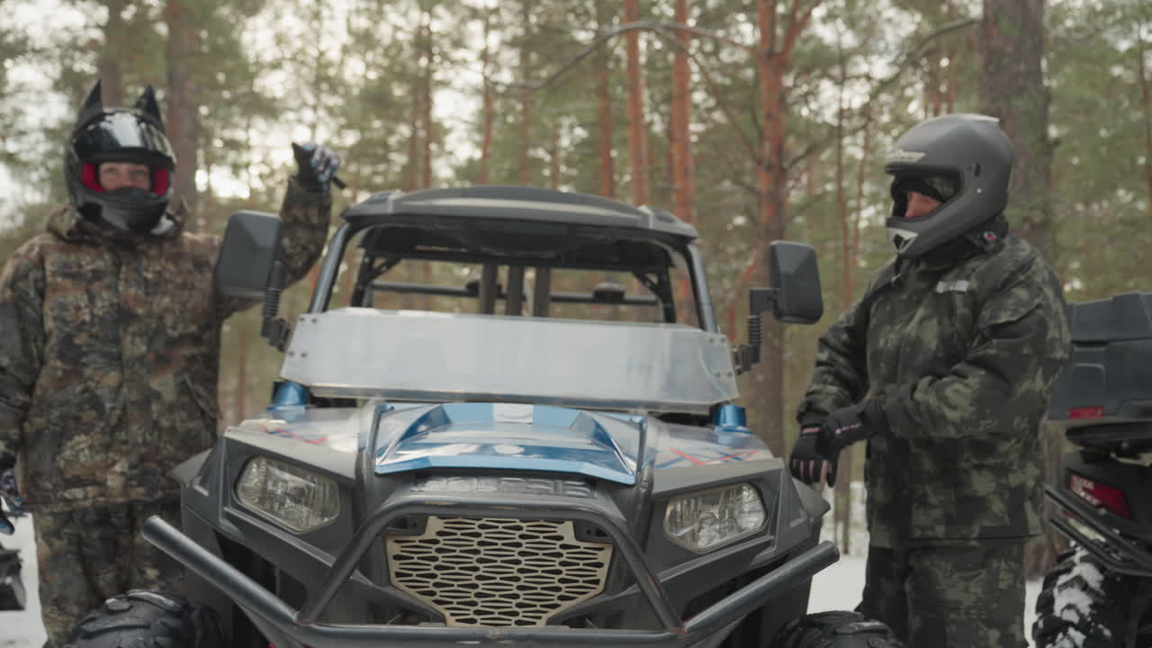 Snowy forest atv convoy with drivers preparing utv. Camouflaged riders in helmets stand beside blue utility vehicle and snowmobile, pine trees and fresh snow, rugged tires and grille, winch visible