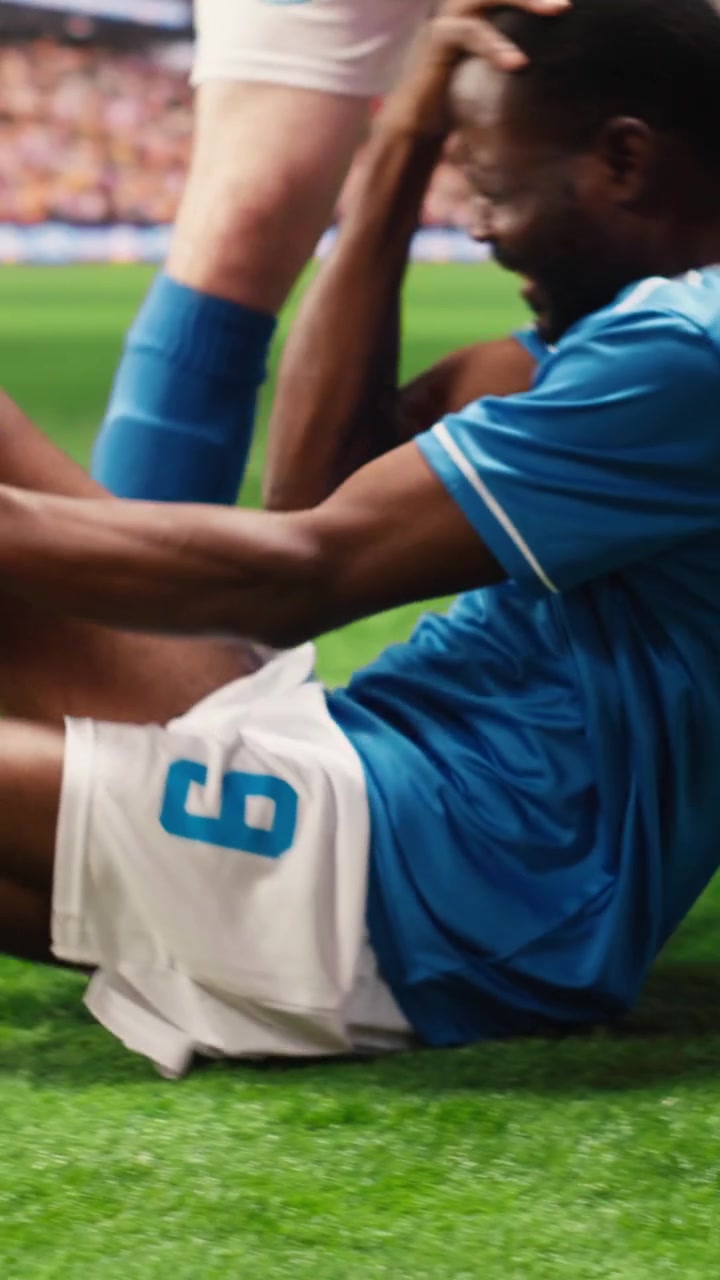 Video verticale: calciatore in maglia blu placcato che rotola sull'erba dopo un infortunio alla gamba.
