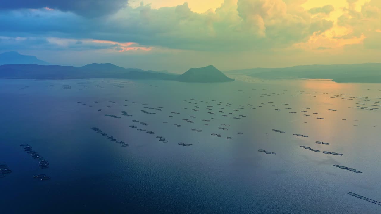 A wide aerial of Taal Volcano with square fishing nets on the lake, glowing sunset contrasting to the deep blue side of dawn in Batangas, Philippines