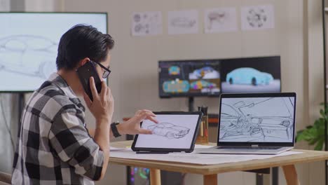 side view of asian male talking on smartphone while drawing new car design concept on a tablet in the studio with tv and computers display 3d electric car model