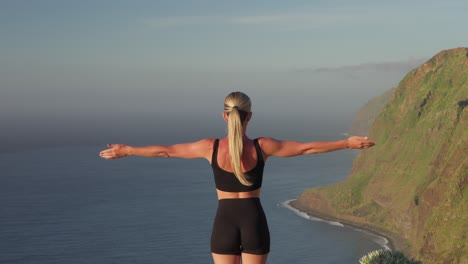 Mujer-De-Yoga-Levantando-Los-Brazos-Hacia-El-Cielo-Mientras-Mira-Los-Acantilados-Costeros.