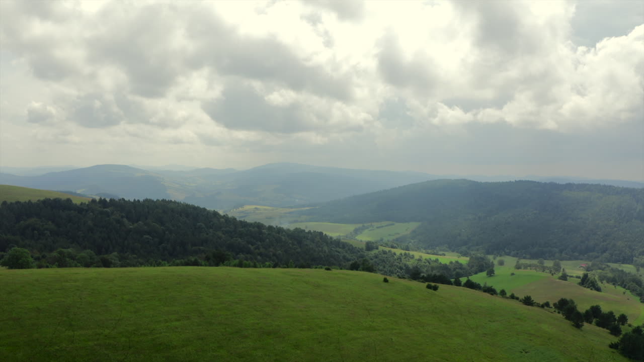 Fast aerial push in panorama of rolling green hills, meadows and forested valleys beneath dramatic clouds - establishing shot. Bieszczady, Poland