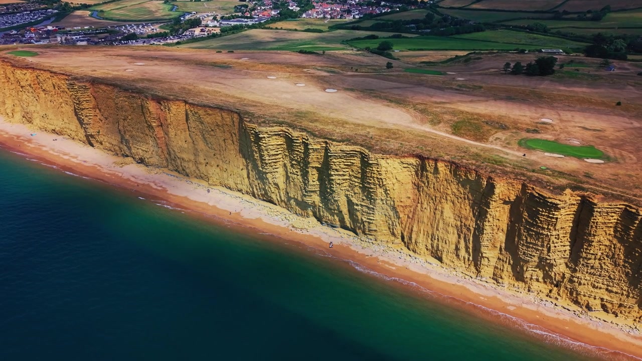 Aerial drone footage revealing the dramatic layered cliff face, narrow beach and turquoise sea at West Bay on Dorset’s Jurassic Coast, England. A coastal golf course and walking paths