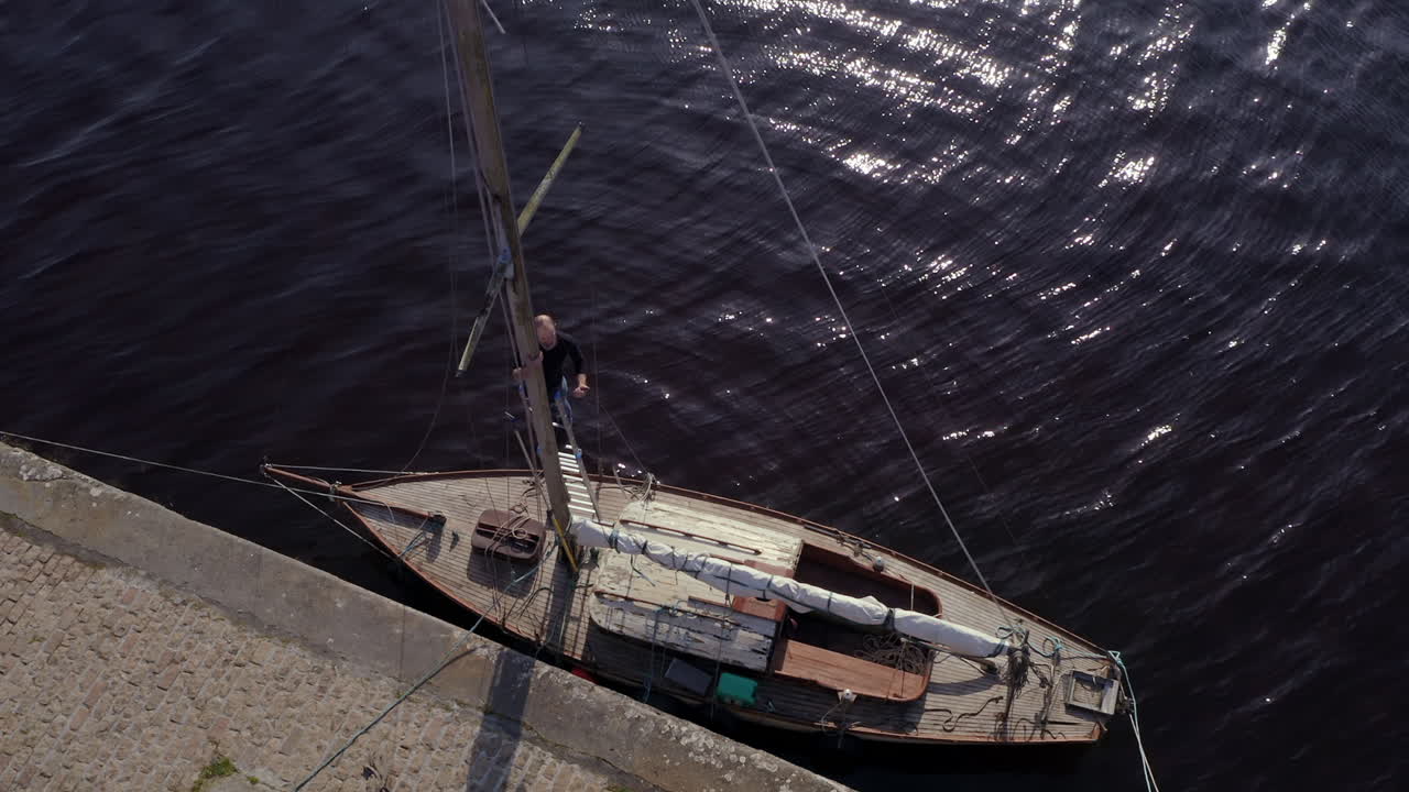 Aerial top down of man balancing on ladder climbing mast of docked boat, calm dark water ripples across pier edge