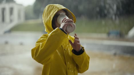 a blonde teenage girl in a yellow jacket drinks water from a white plastic bottle and looks at the heavy rain in the park