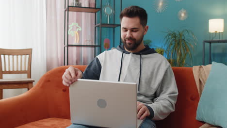 Young-man-freelancer-sitting-on-couch-closing-laptop-pc-after-finishing-work-in-living-room-at-home