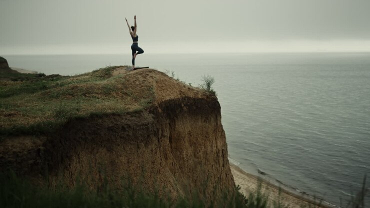 Girl doing yoga tree pose on beach hilltop. Woman standing on one leg outdoor